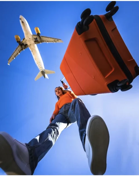 Una viaggiatrice con una valigia arancione accanto a un aereo sotto un cielo azzurro